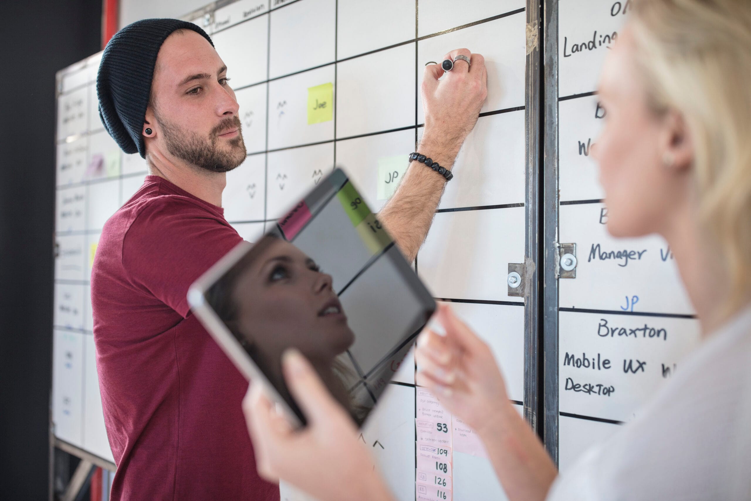 A man writes on a whiteboard while a woman holds a tablet. A man writes on a whiteboard while a woman holds a tablet.